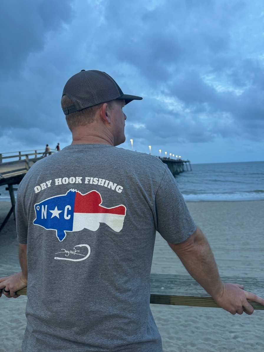 Man wearing a gray t-shirt with a fishing graphic and text, standing on a beach with a pier and ocean view.